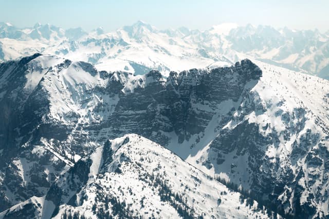 Jagged snow-covered mountain ridge rising amid a vast alpine range under a pale blue sky