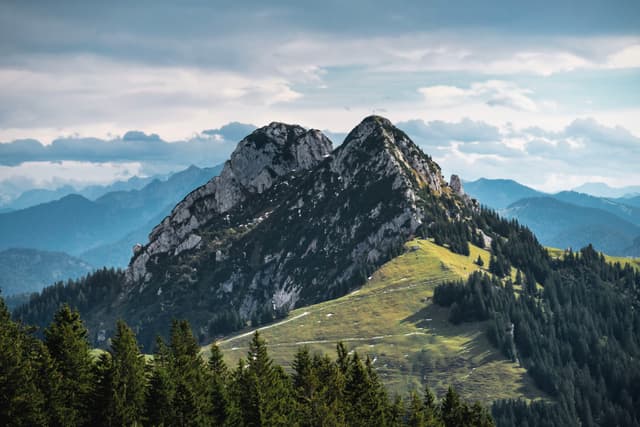 Twin rocky mountain peaks rising above a forested ridgeline with rolling hills and distant hazy ranges under a cloudy sky