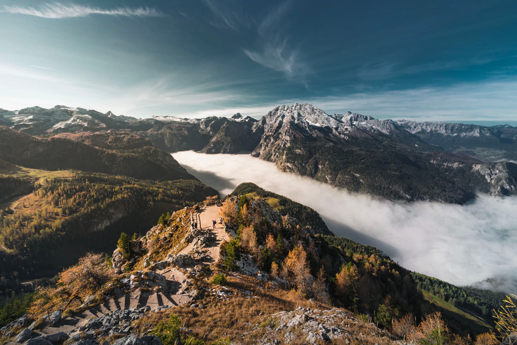 High mountain ridge overlooking a valley filled with a dense layer of clouds beneath snow-capped peaks under a clear blue sky