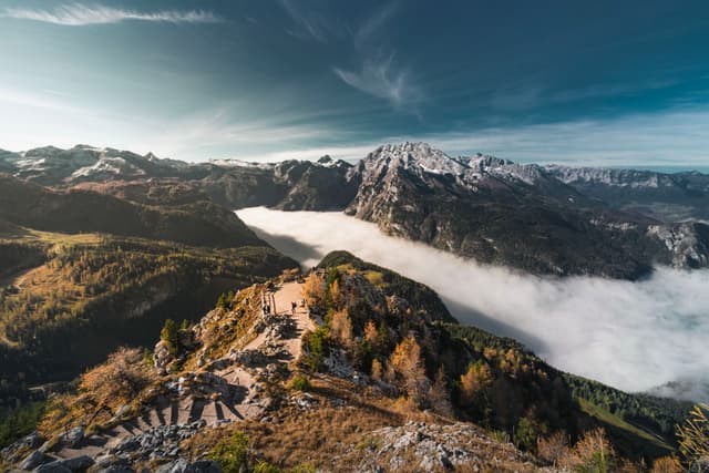 High mountain ridge overlooking a valley filled with a dense layer of clouds beneath snow-capped peaks under a clear blue sky
