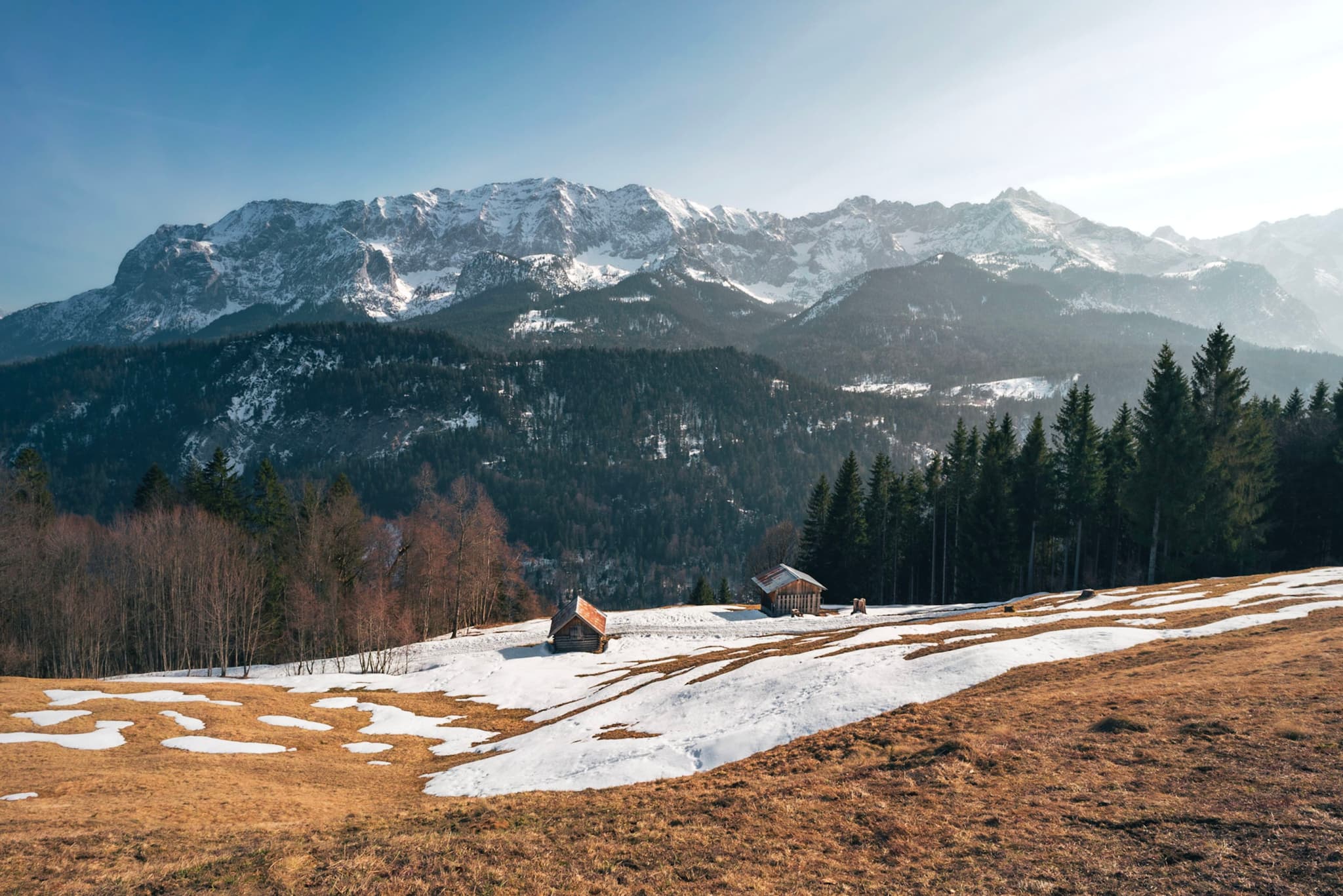 A scenic landscape with snow-dusted mountains in the background, a mix of trees and open fields, and a small cabin in the foreground