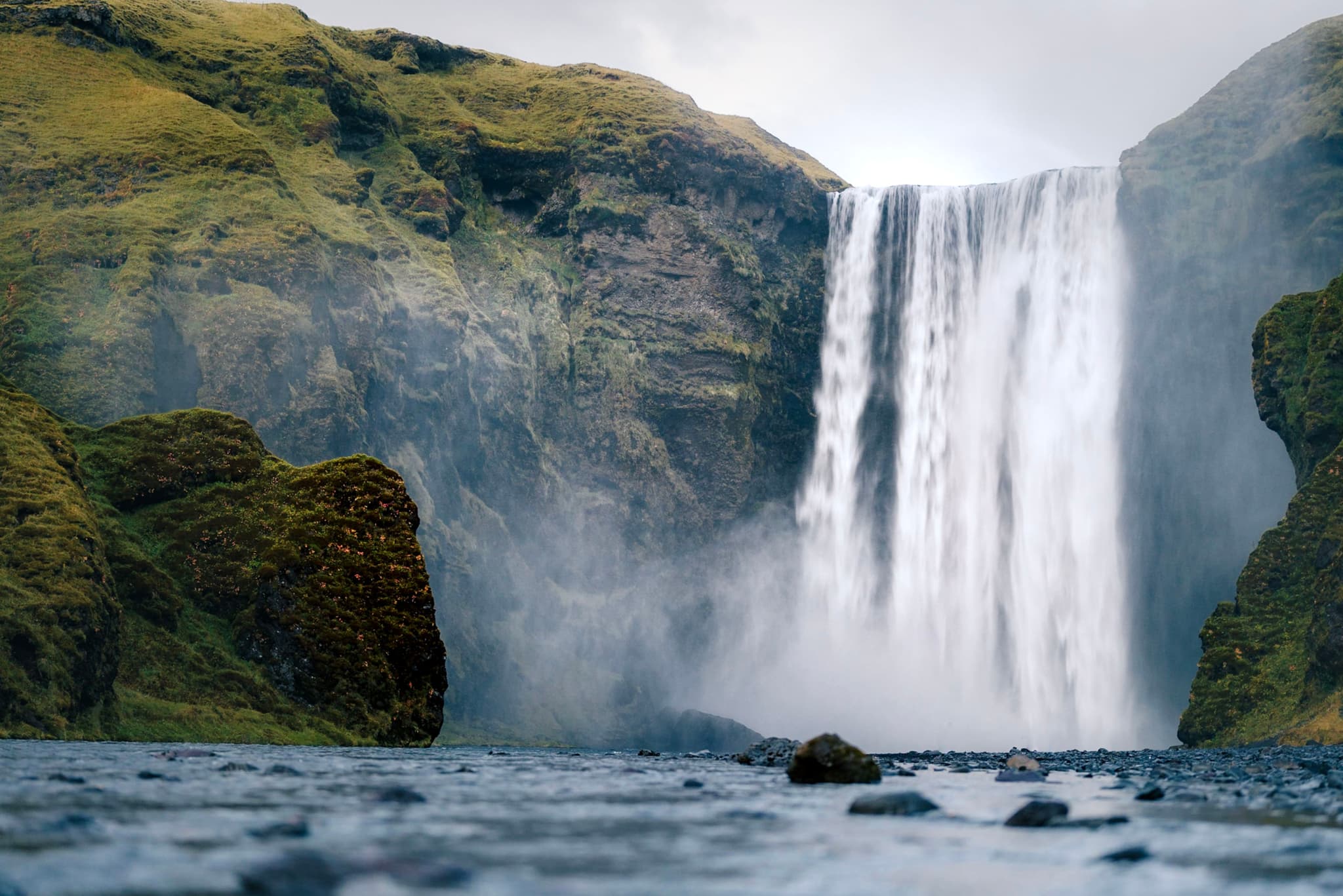 Powerful waterfall plunging over a mossy cliff into a rocky river beneath overcast skies
