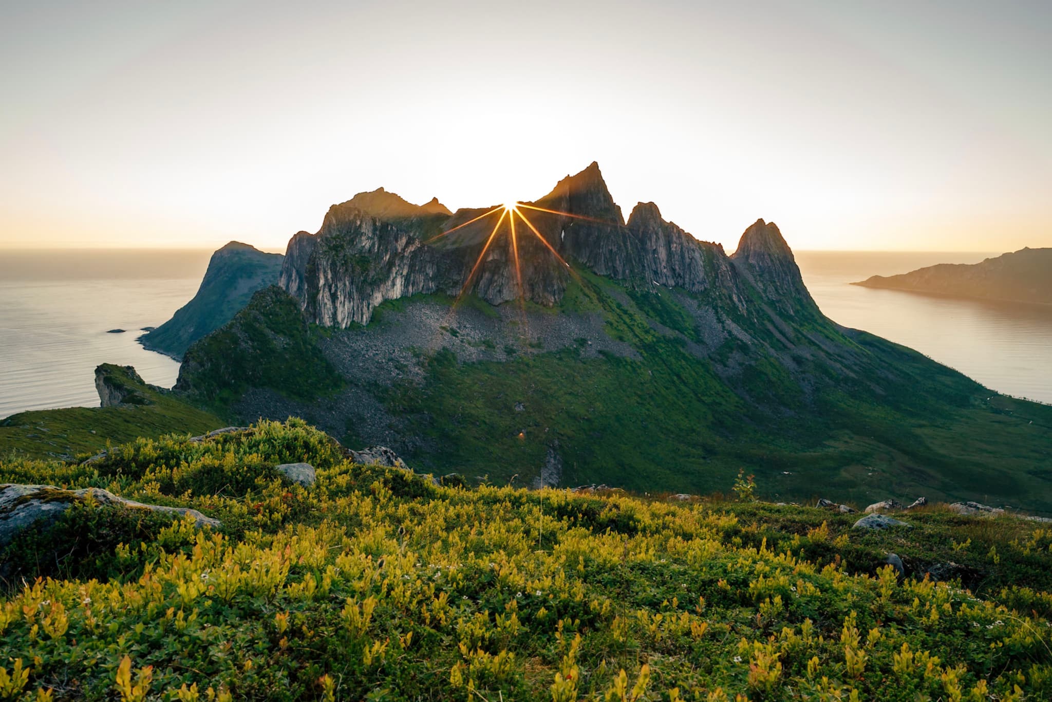 A mountain range with jagged peaks is illuminated by the sun setting or rising behind it, surrounded by lush greenery and water