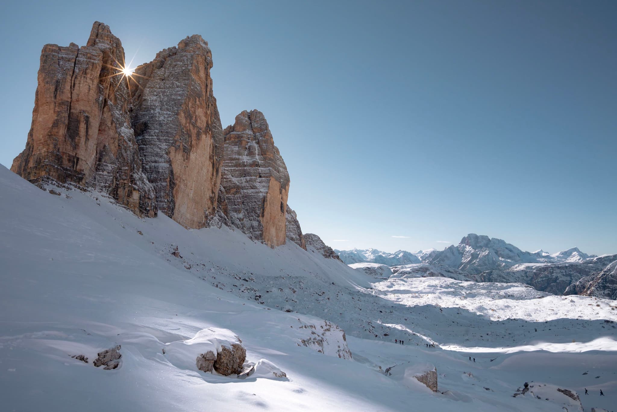 Towering rocky peaks rise from a snow-covered alpine landscape under a clear blue sky, with distant mountain ranges stretching toward the horizon