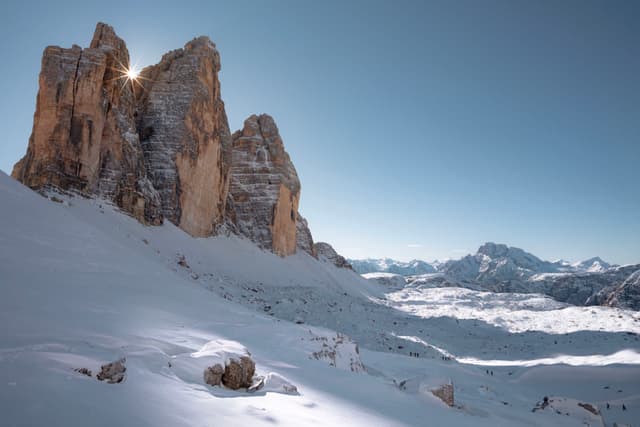 Towering rocky peaks rise from a snow-covered alpine landscape under a clear blue sky, with distant mountain ranges stretching toward the horizon