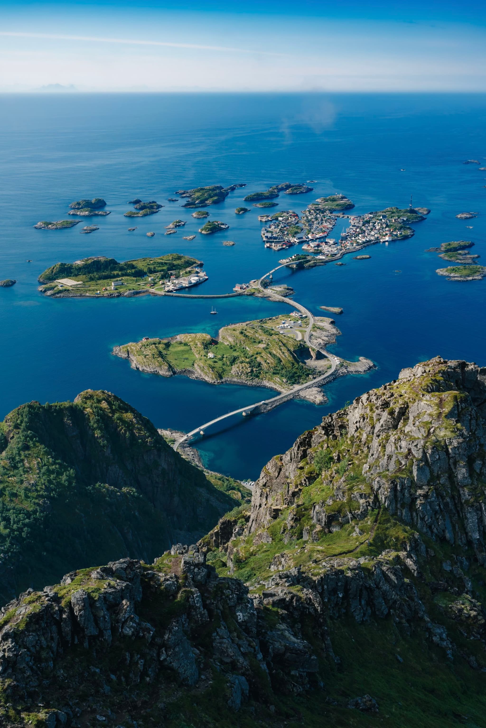 Aerial view of a coastal landscape with rugged mountains in the foreground, a bridge connecting small islands, and a vast expanse of blue ocean in the background