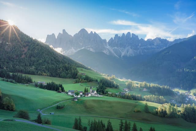 A picturesque valley with lush green fields, a small village, and a backdrop of majestic mountains under a clear blue sky with the sun peeking over the horizon