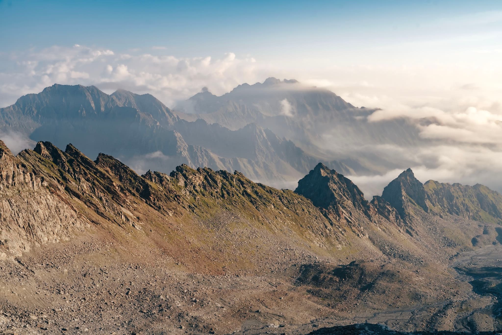 A rugged mountain range with jagged peaks under a clear sky, partially covered by clouds