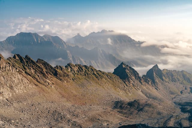 A rugged mountain range with jagged peaks under a clear sky, partially covered by clouds