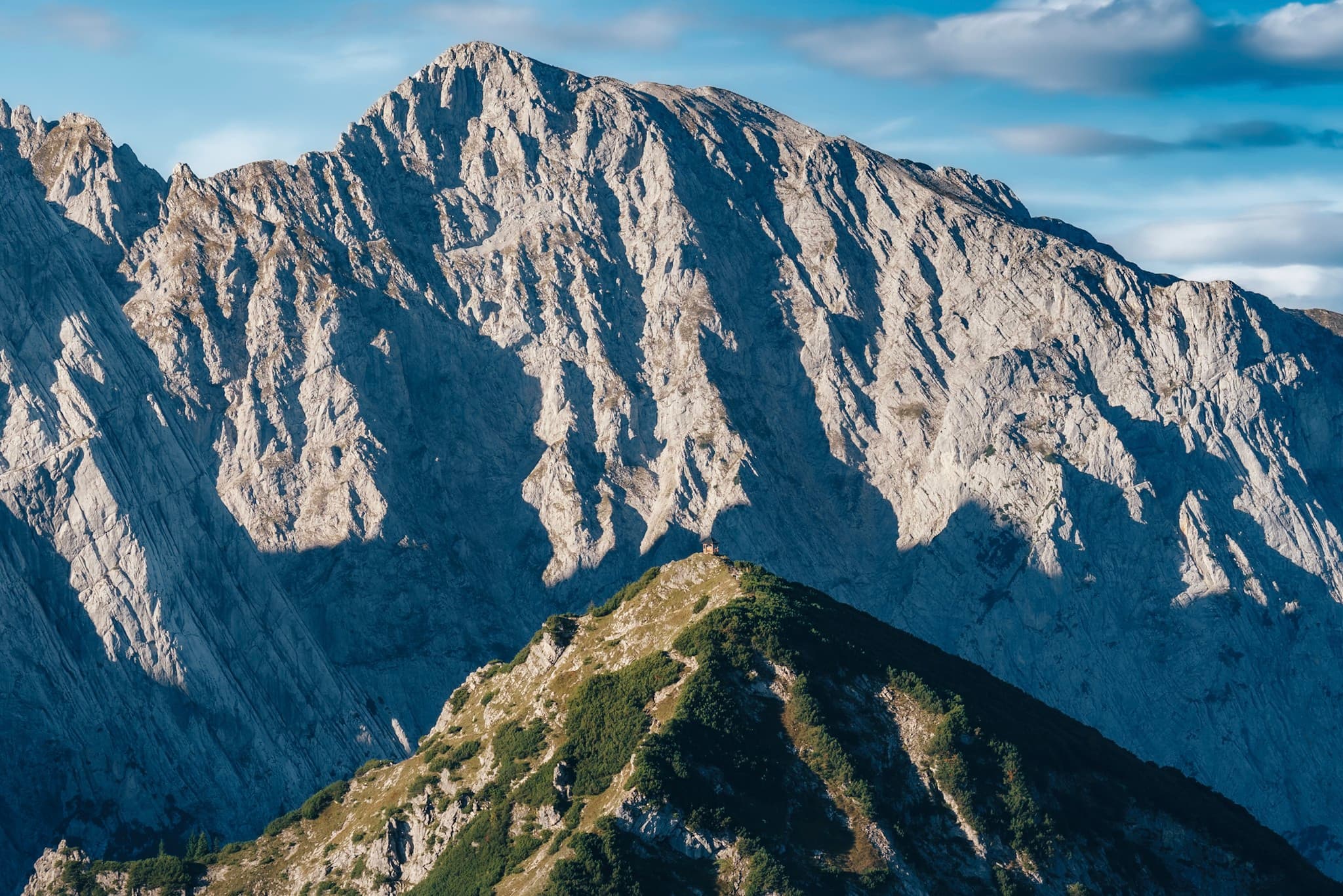 Jagged rocky massif rises behind a green, narrow mountain ridge under a blue sky with scattered clouds