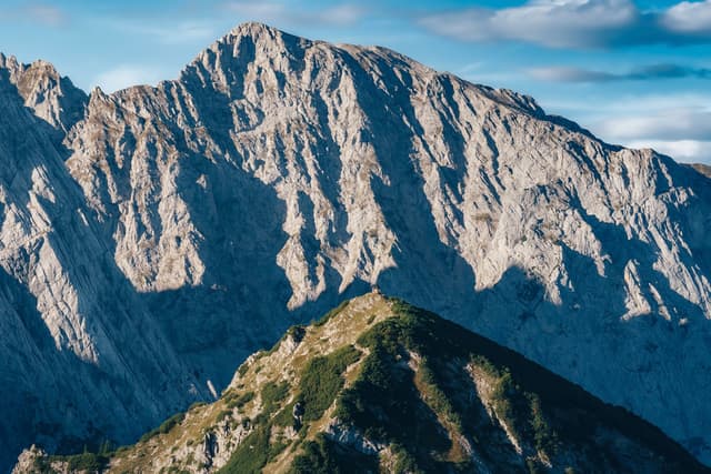 Jagged rocky massif rises behind a green, narrow mountain ridge under a blue sky with scattered clouds