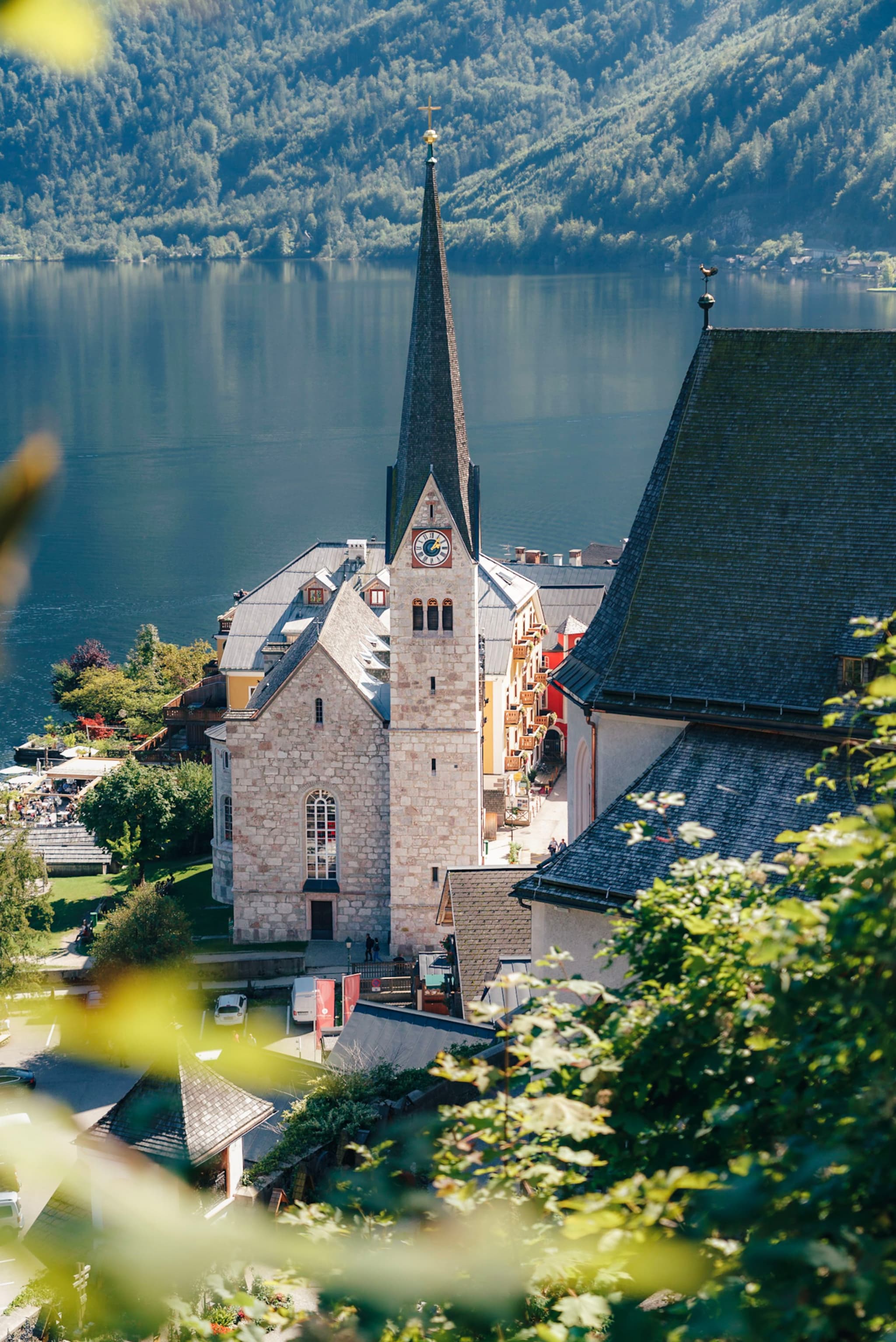 A picturesque village with a prominent church featuring a tall spire, set against a backdrop of a serene lake and lush, forested mountains