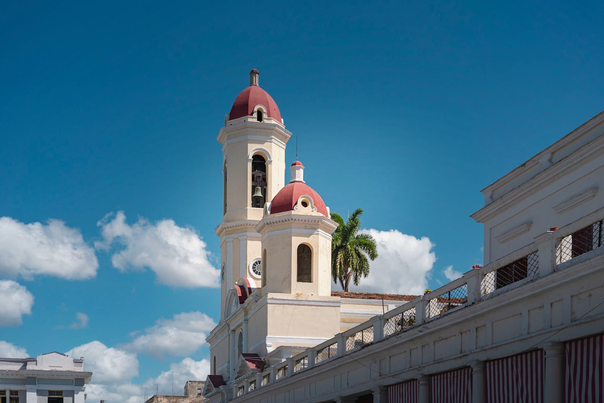 White church with twin red-domed towers rising above surrounding buildings under a bright blue sky with scattered clouds