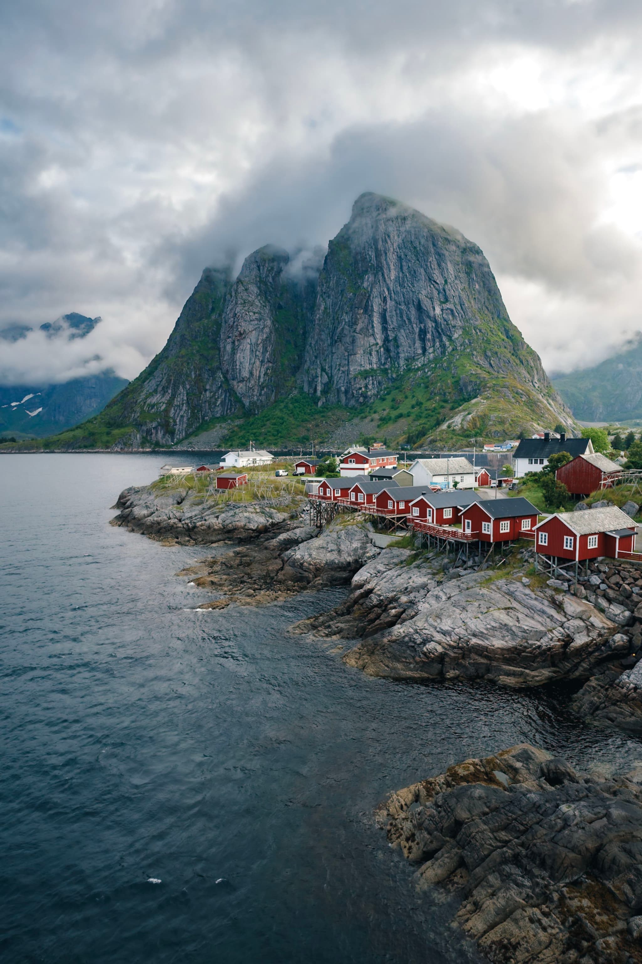 A coastal village with red houses sits on rocky shores, backed by a towering, cloud-covered mountain