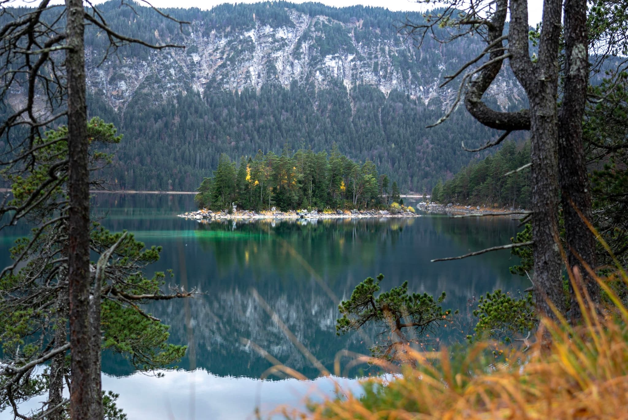 A serene lake with a small, tree-covered island in the center, surrounded by forested mountains. The water reflects the landscape, and the scene is framed by trees in the foreground