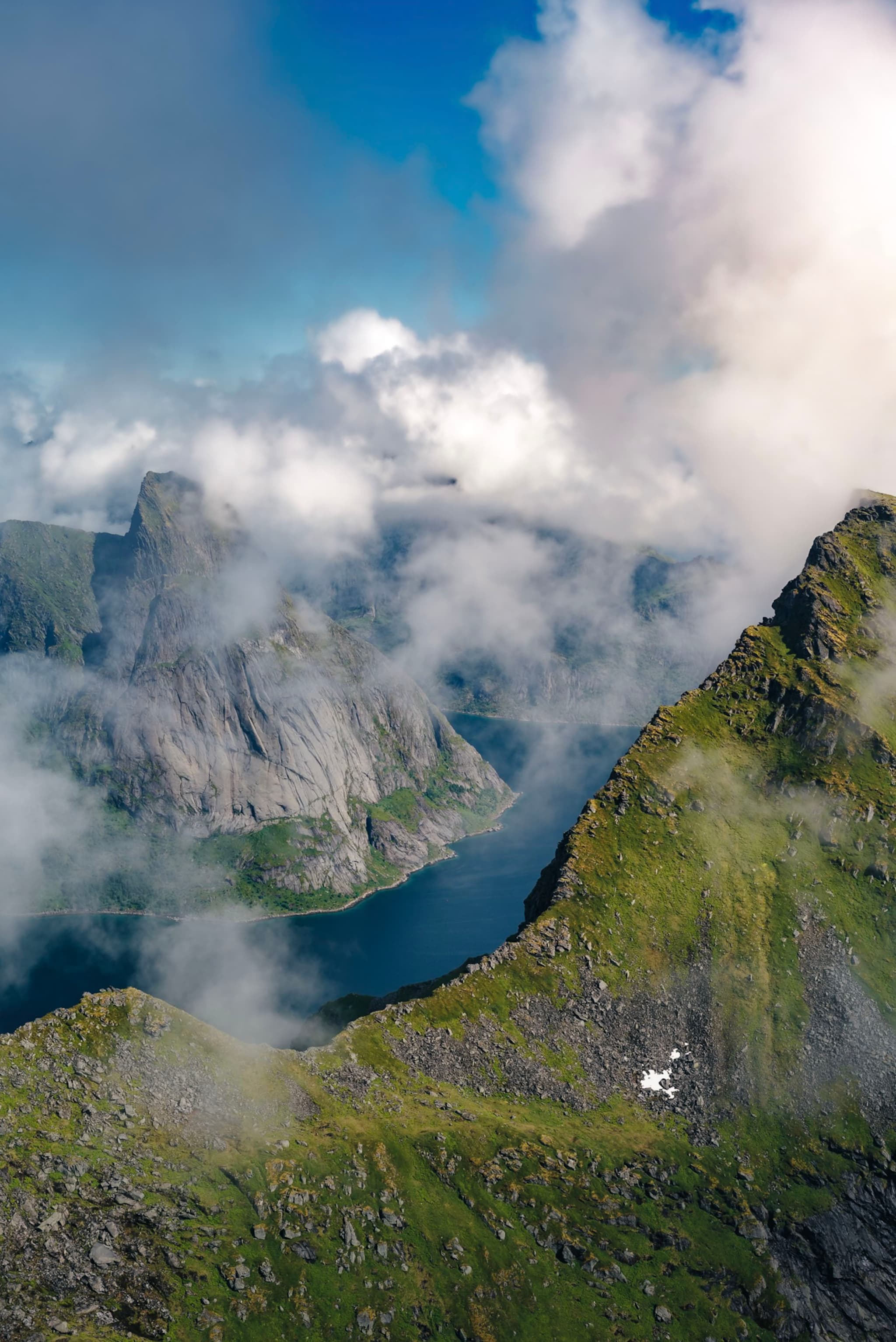 Aerial view of a mountainous landscape with lush green peaks and a winding body of water, partially obscured by clouds