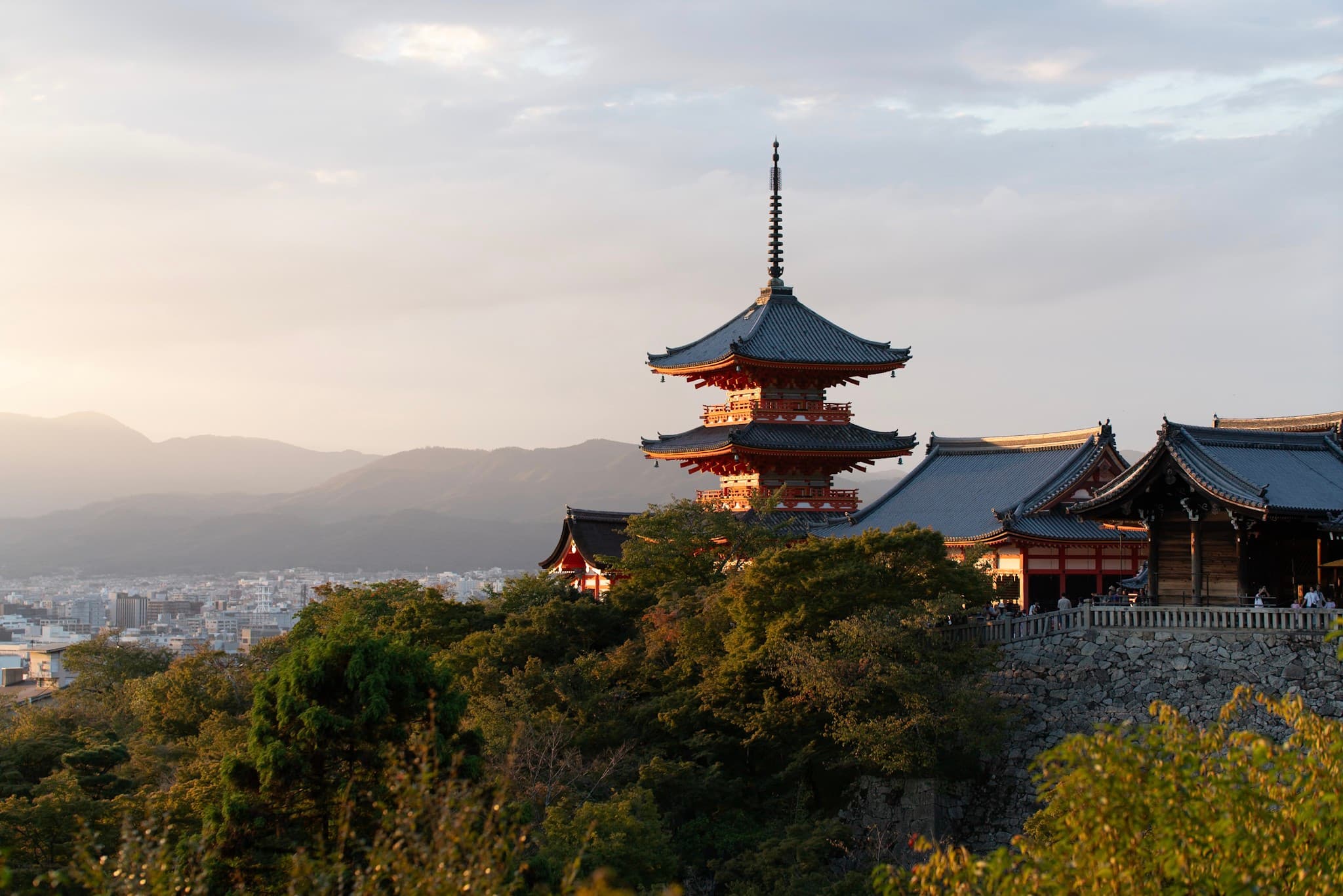 Traditional Japanese pagoda on a forested hillside overlooking a distant city at sunset