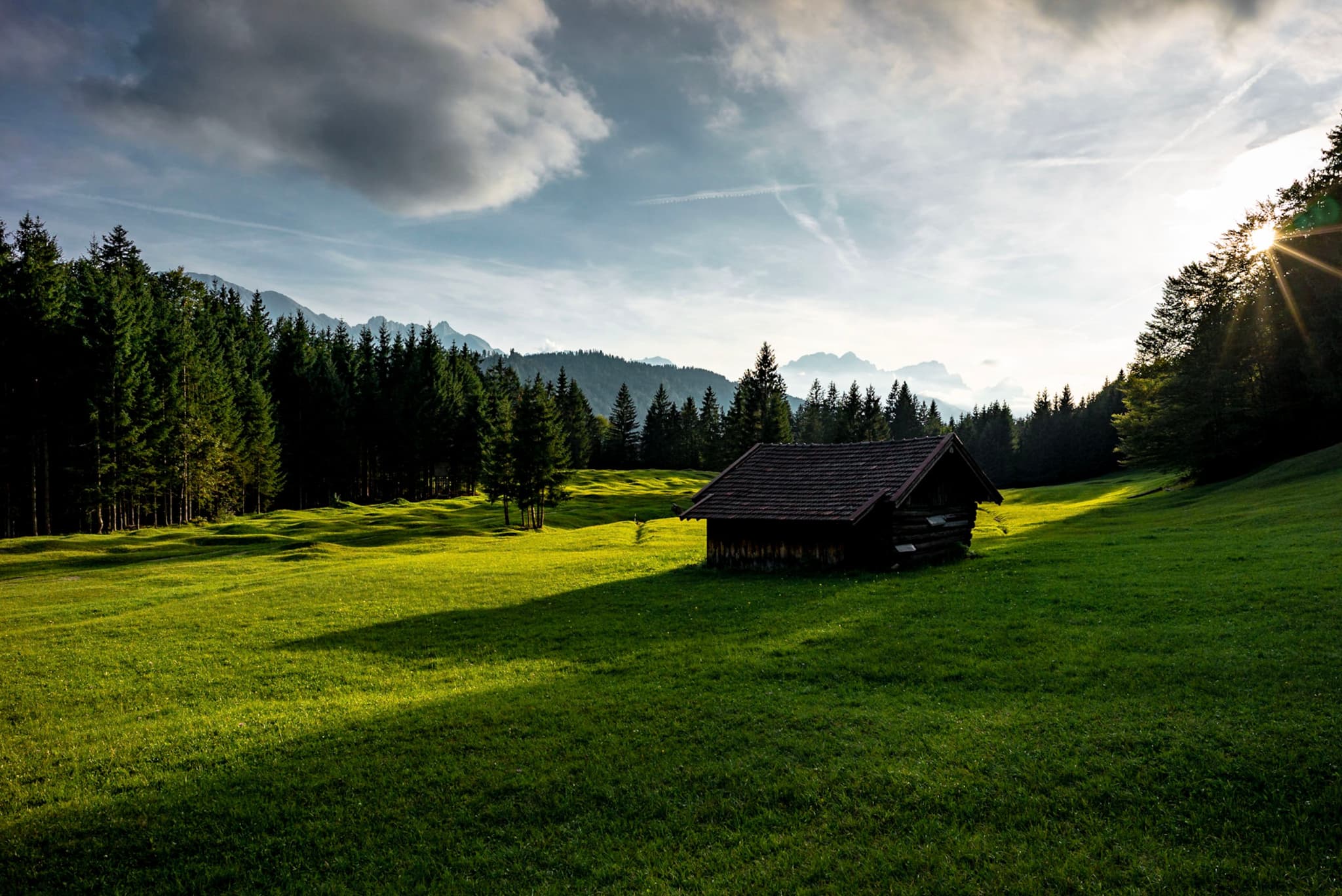 Small dark cabin standing in a sunlit green meadow bordered by dense evergreen forest and distant mountains under a partly cloudy sky with low sun rays