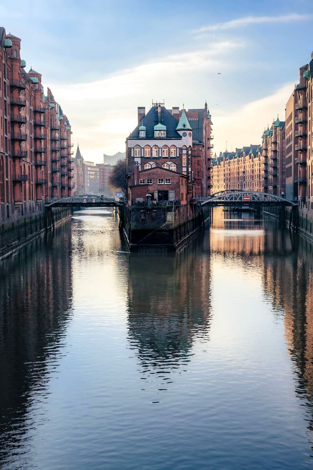 Historic brick buildings lining a calm canal with a central house on a narrow island and small bridges connecting both sides