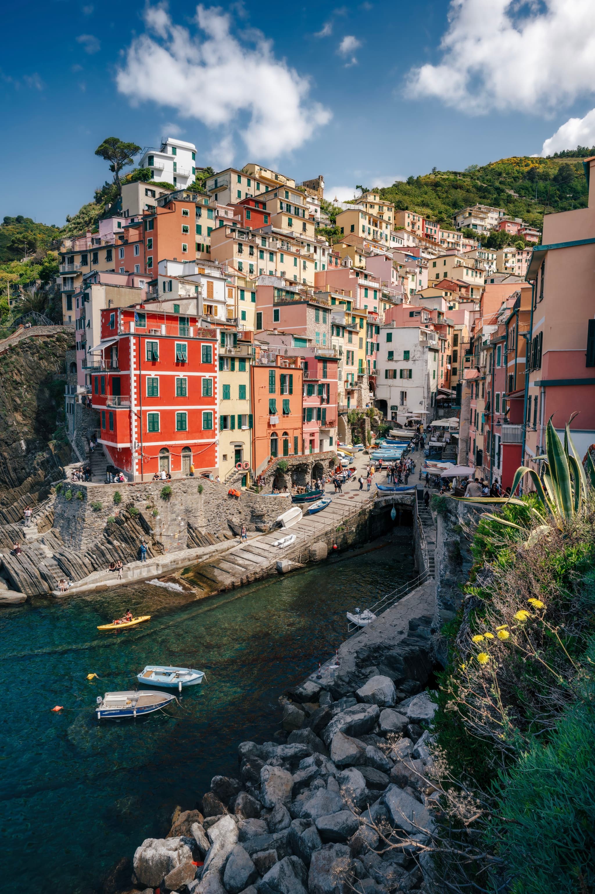 A coastal village with colorful buildings perched on a hillside, overlooking a small harbor with boats, surrounded by rocky terrain and clear blue water
