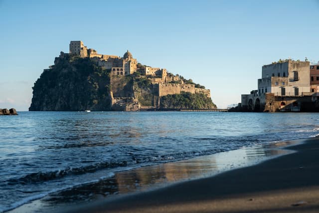 Ancient stone castle complex perched on a rocky island just offshore, with calm waves lapping a sandy beach and a small coastal town building on the right under a clear blue sky