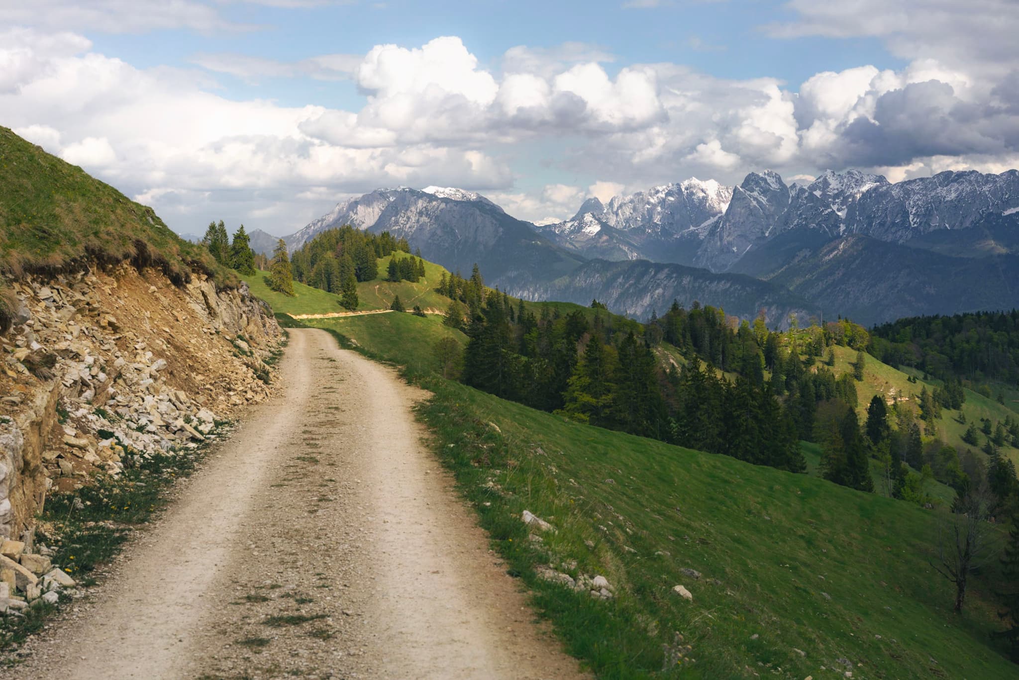 Winding dirt trail cutting through green alpine hillsides toward distant snow-capped mountains under a partly cloudy sky