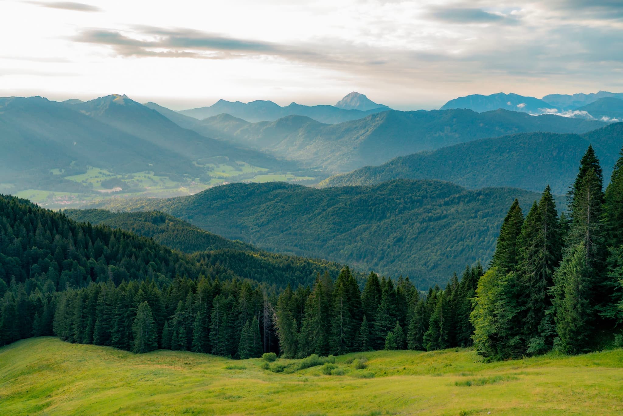 Layered mountain ranges with sunbeams breaking through clouds, illuminating green valleys and dense evergreen trees in the foreground