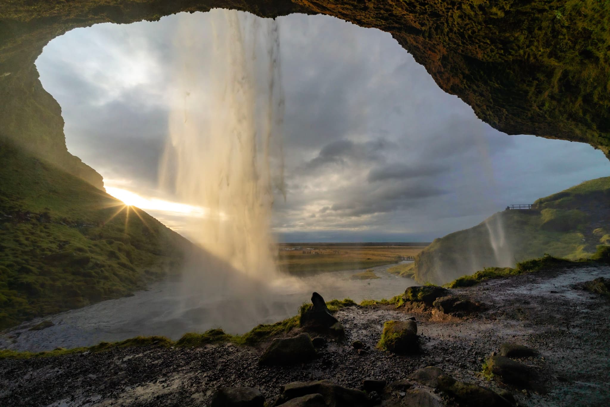 View from behind a tall waterfall cascading over a rocky overhang with sun rays breaking through the mist onto the landscape below
