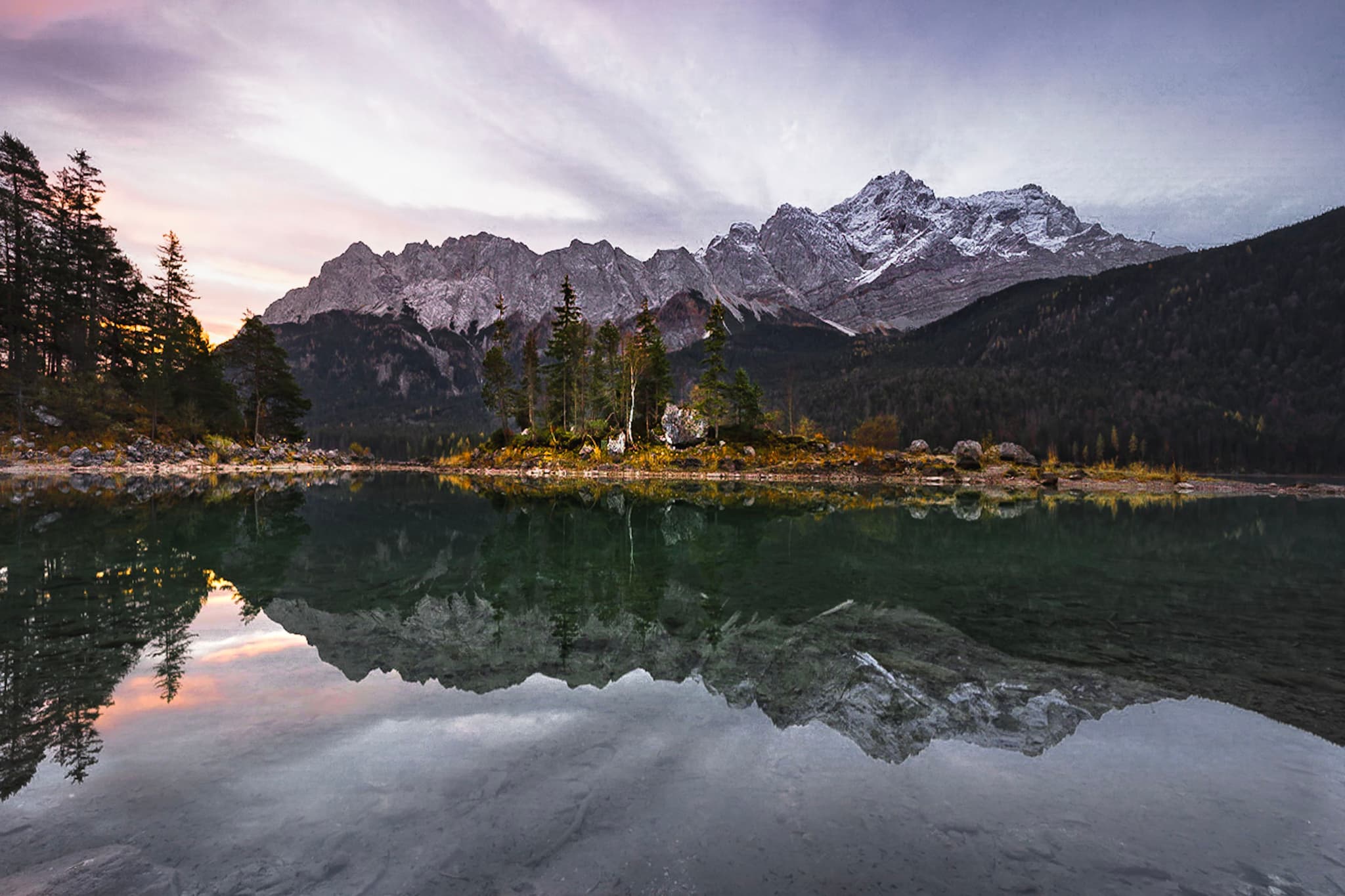 A serene mountain landscape with snow-capped peaks in the background, a small island with trees in the middle ground, and a calm lake reflecting the scene