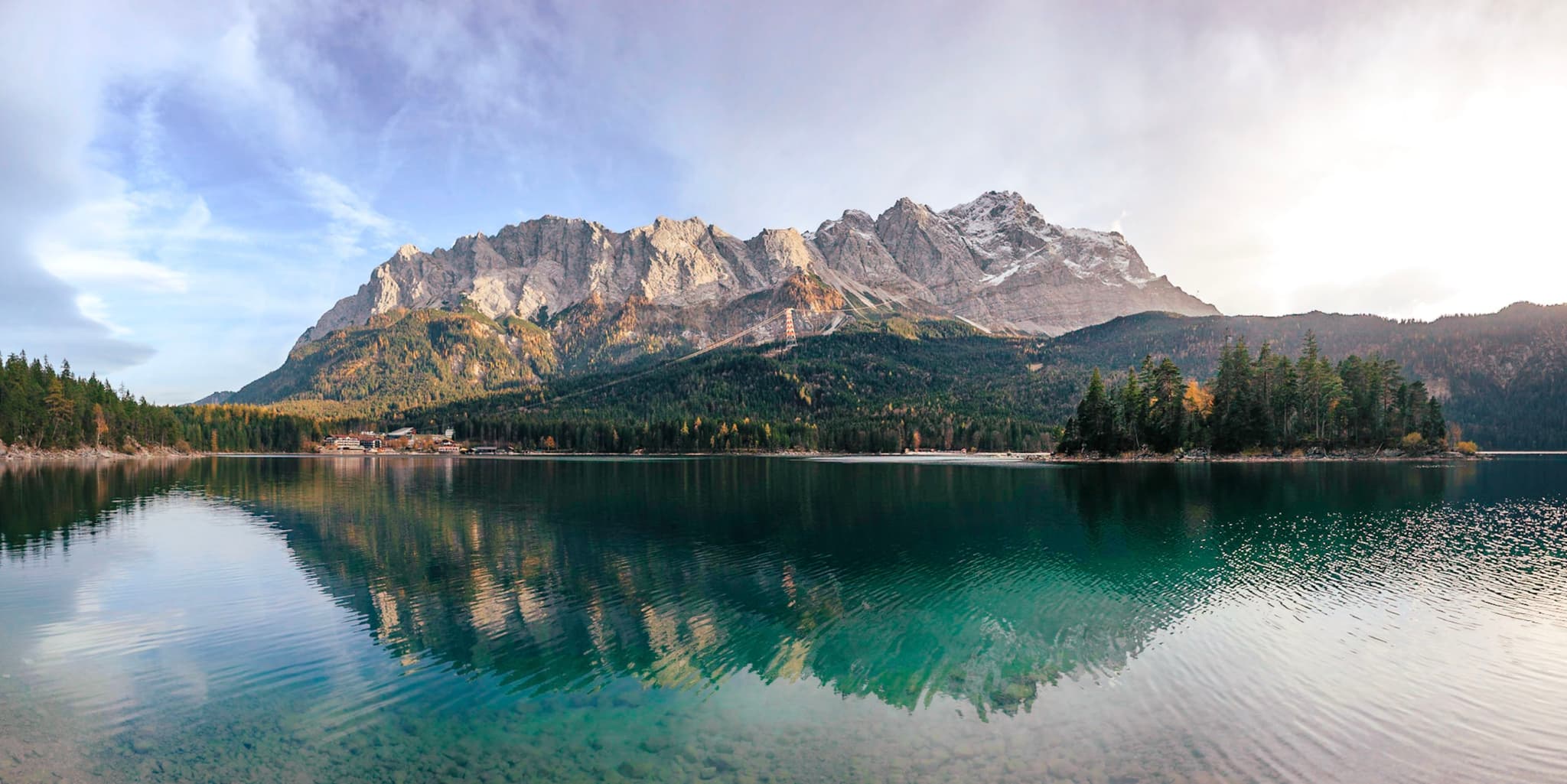 A serene lake with clear turquoise water reflects a majestic mountain range under a partly cloudy sky, surrounded by lush green forests