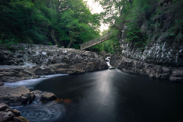 A serene river flows through a rocky gorge, surrounded by lush green trees. A wooden bridge spans across the gorge, and the water appears smooth and reflective