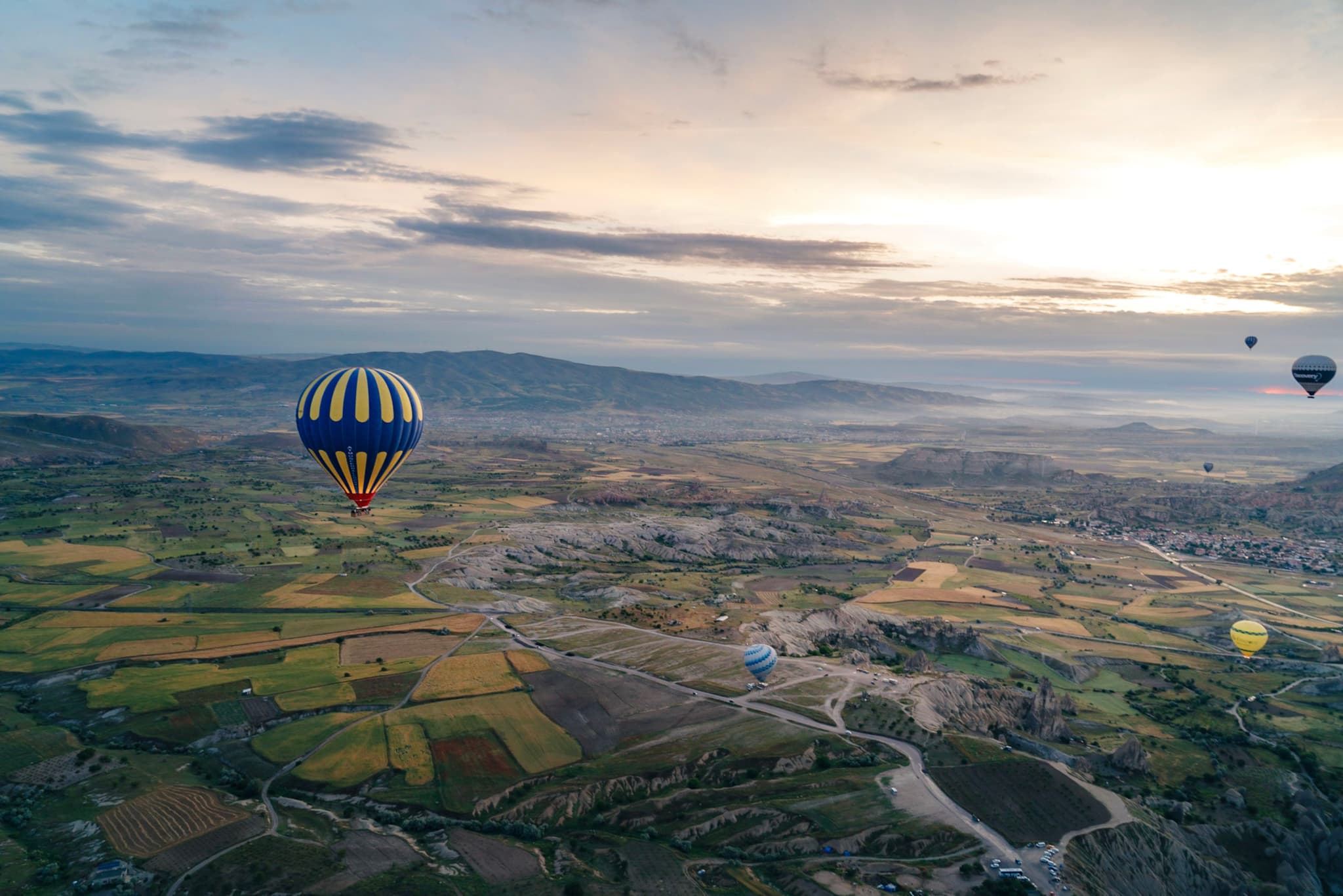 Hot air balloons floating over a vast, patchwork landscape at sunrise, with distant mountains and a partly cloudy sky