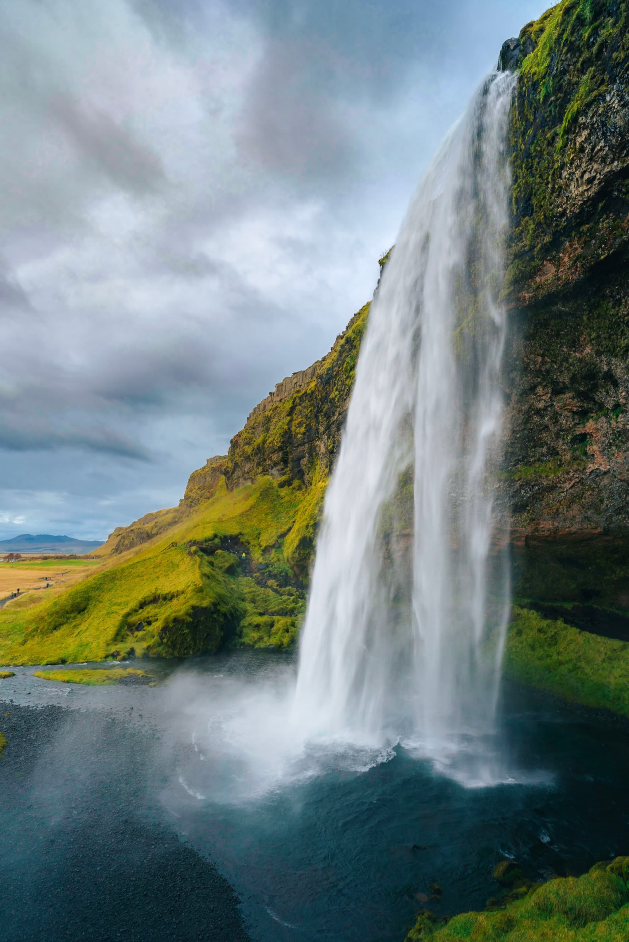 Tall waterfall plunging from a mossy cliff into a dark pool under a cloudy sky