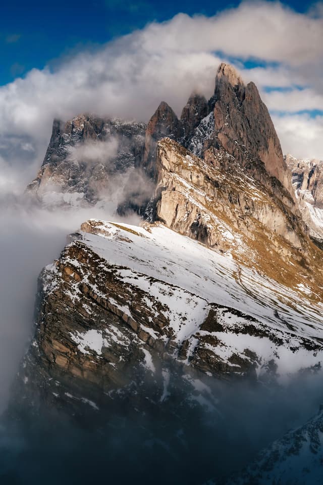 A dramatic mountain peak rises through clouds, with snow-covered slopes and rugged rock formations under a blue sky