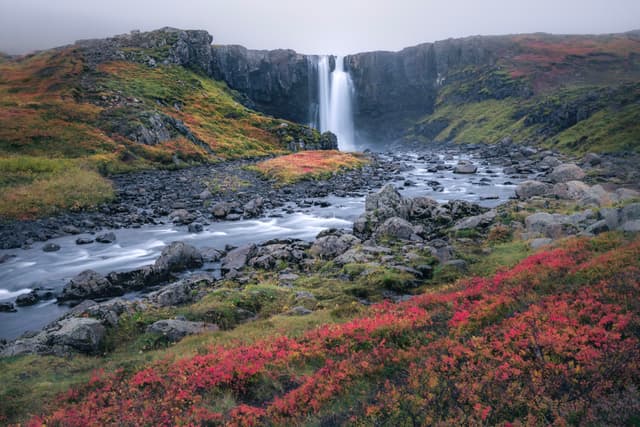 Waterfall pouring over a rocky cliff into a winding river, surrounded by autumn foliage in red and orange hues under an overcast sky