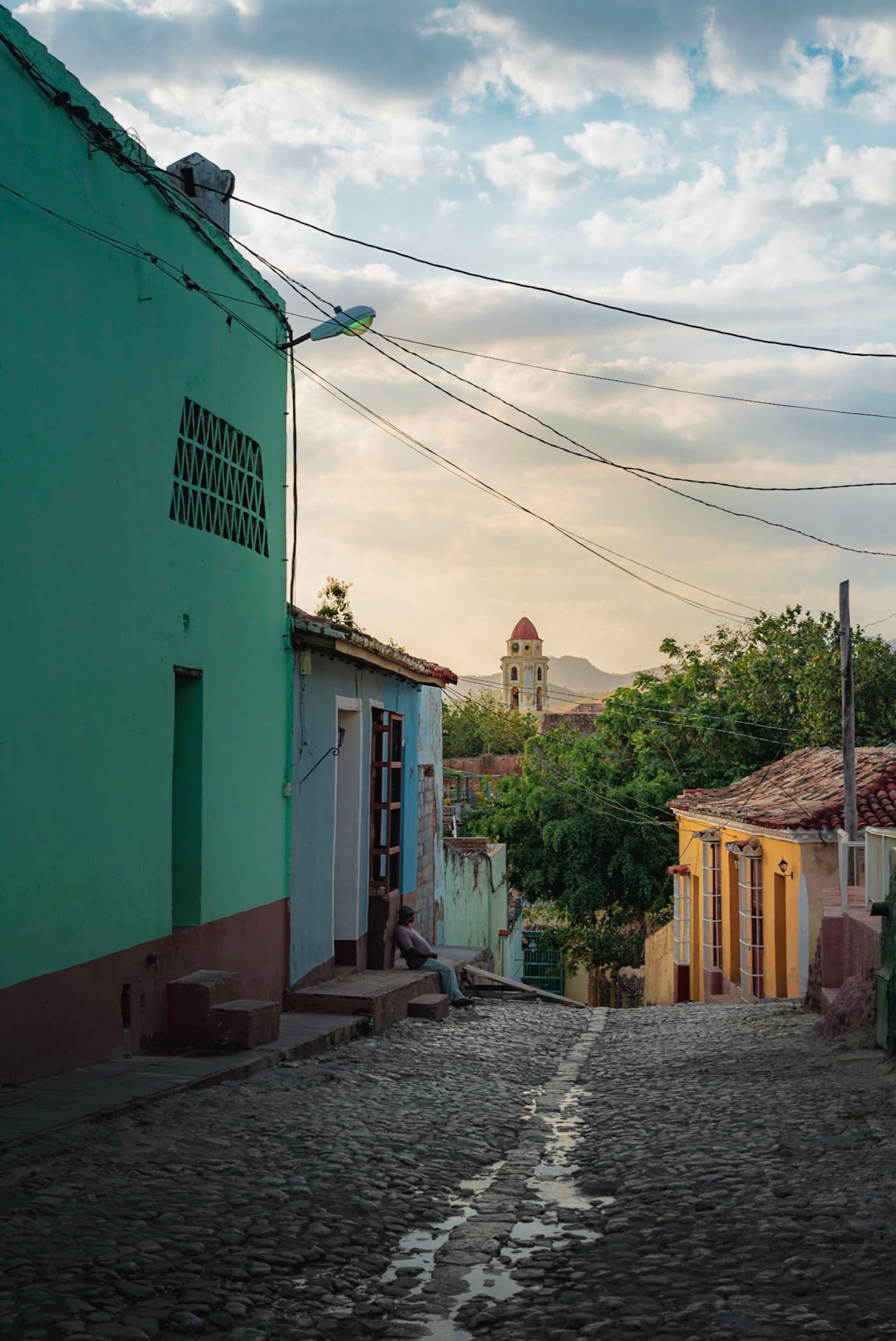 Narrow cobblestone street lined with colorful houses descending toward a distant bell tower at sunset