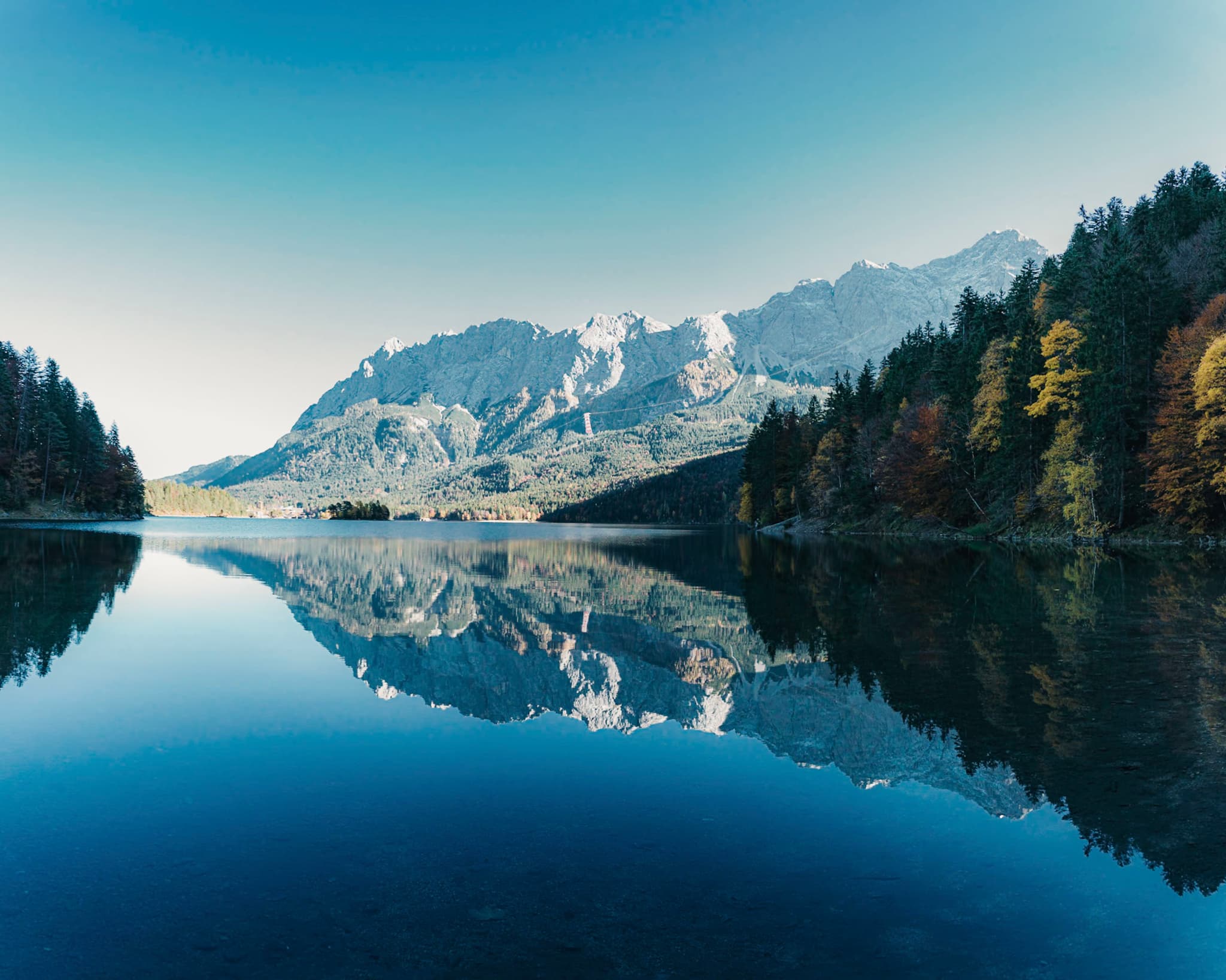 A serene lake reflects surrounding mountains and trees under a clear blue sky