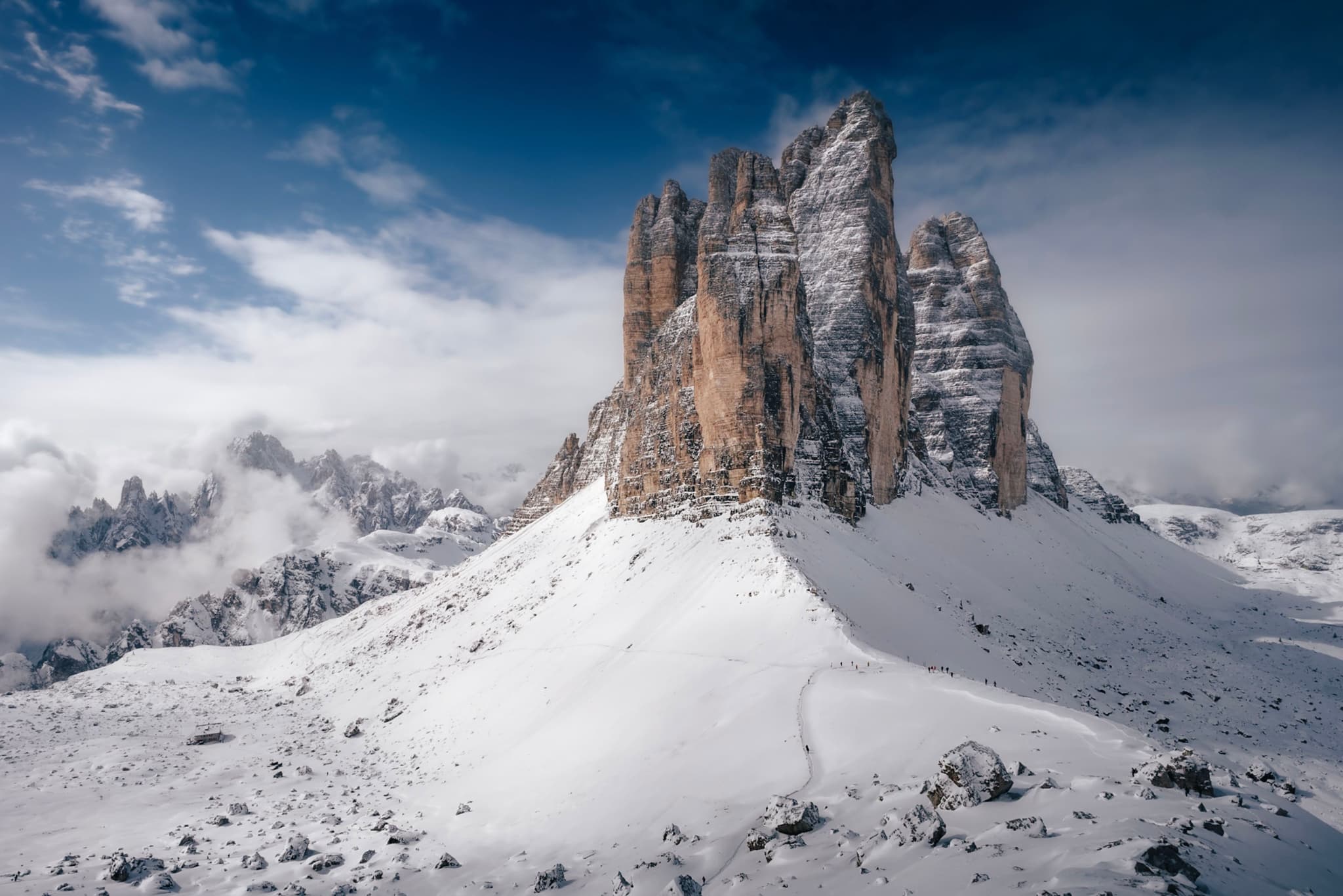 A snow-covered mountain peak under a partly cloudy sky, with rugged rock formations and distant mountains in the background