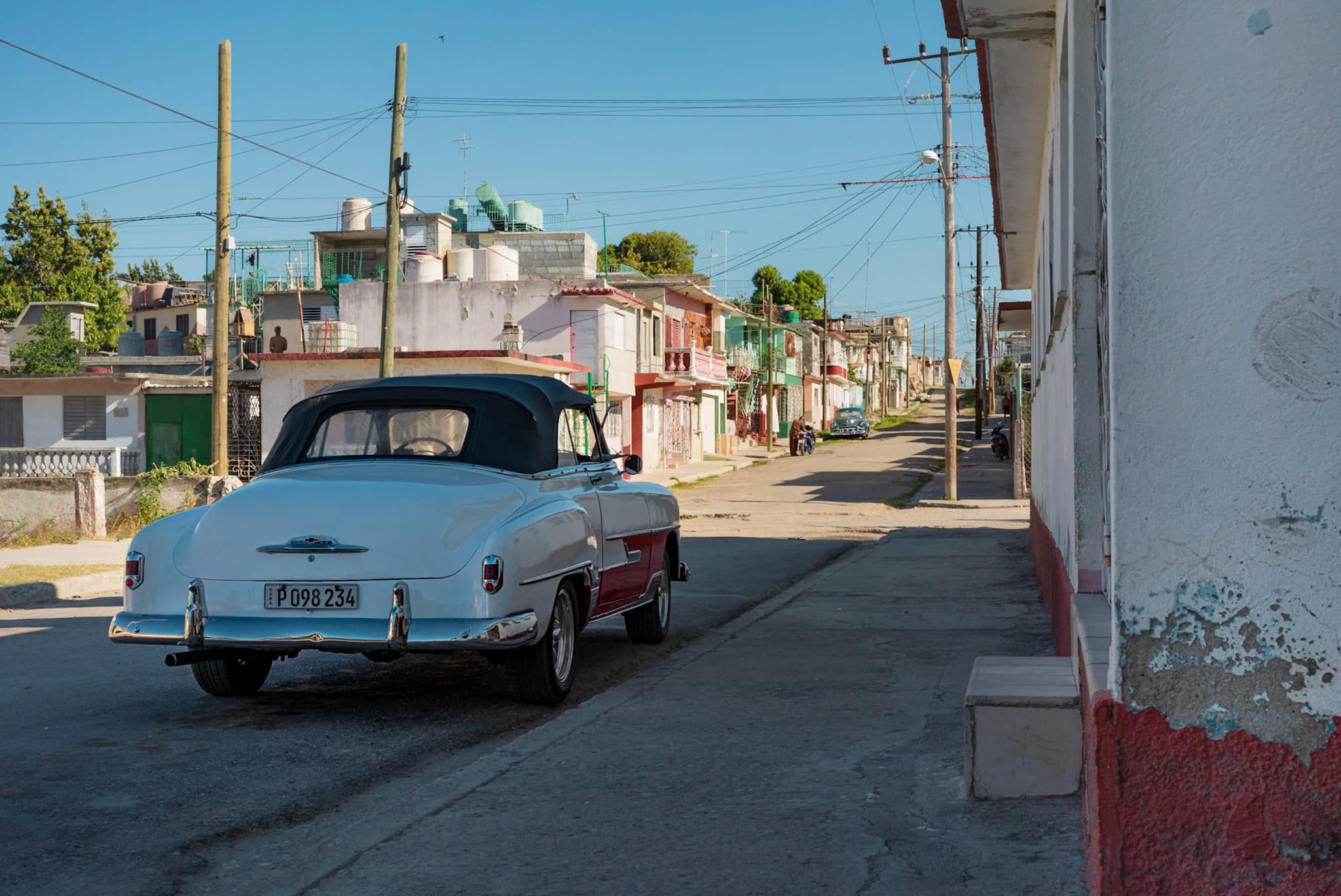 Classic vintage car parked on a quiet sunlit street lined with modest houses and utility poles in a small town neighborhood