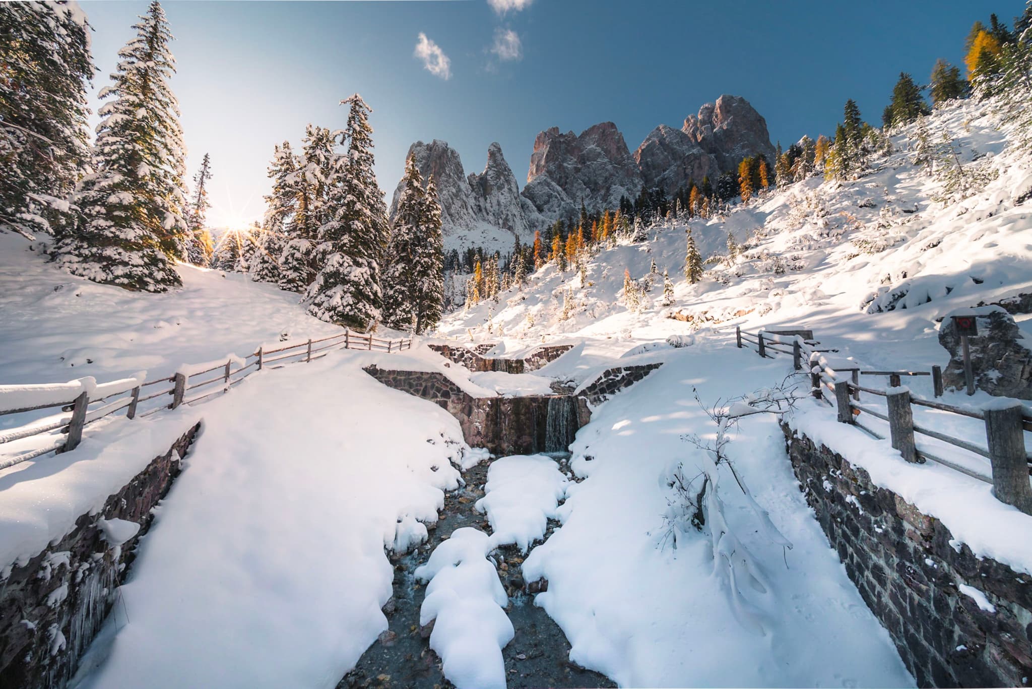 Snow-covered mountain pass with fenced stone walls and a frozen stream leading toward jagged peaks under a clear blue sky