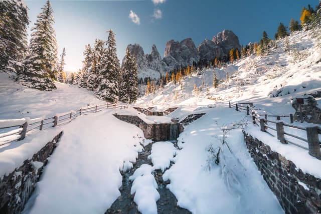 Snow-covered mountain pass with fenced stone walls and a frozen stream leading toward jagged peaks under a clear blue sky