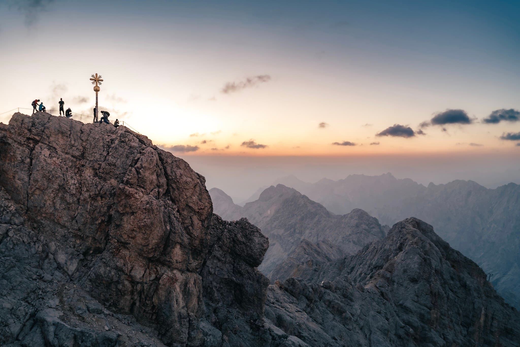 Silhouetted hikers stand by a summit cross on a rugged mountain peak at sunrise, overlooking layers of distant ridges under a soft, colorful sky