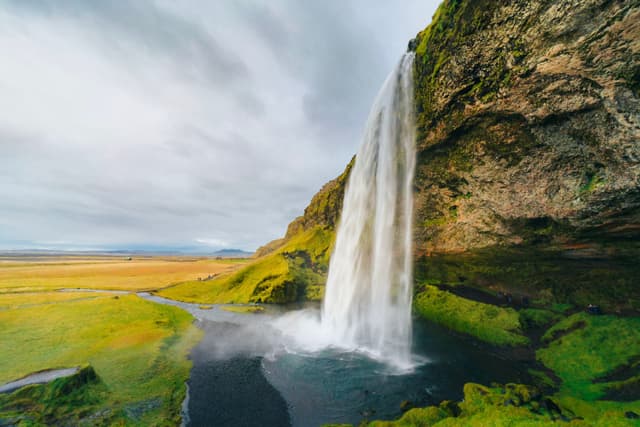 Tall waterfall plunging from a mossy cliff into a winding stream across open plains under an overcast sky