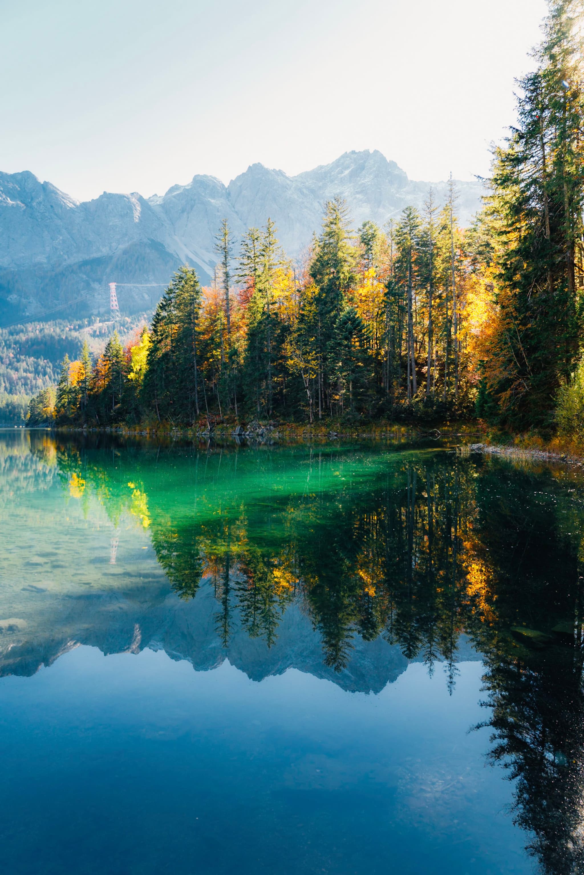 Clear alpine lake reflecting a line of autumn-colored evergreen trees with distant mountain peaks under a bright sky