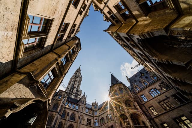 Upward view from a stone courtyard framed by tall historic buildings with a central clock tower and sunburst in the clear sky