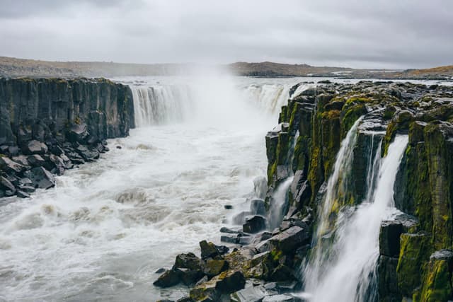 Powerful white water river crashing through rocky cliffs with multiple waterfalls under an overcast sky