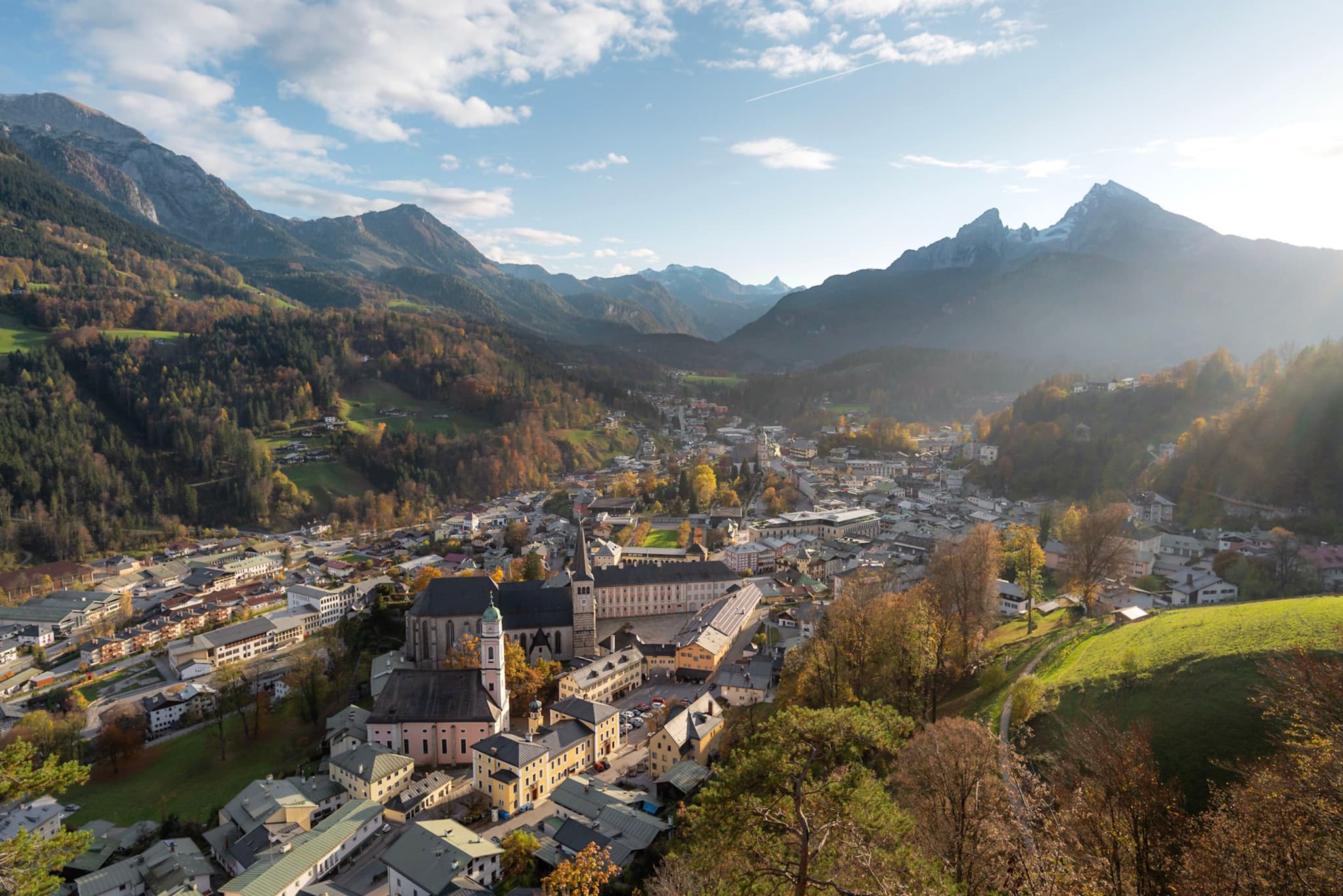 A sunlit mountain valley village with clustered houses, a prominent church, and surrounding forested slopes under a partly cloudy sky