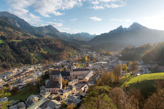 A sunlit mountain valley village with clustered houses, a prominent church, and surrounding forested slopes under a partly cloudy sky