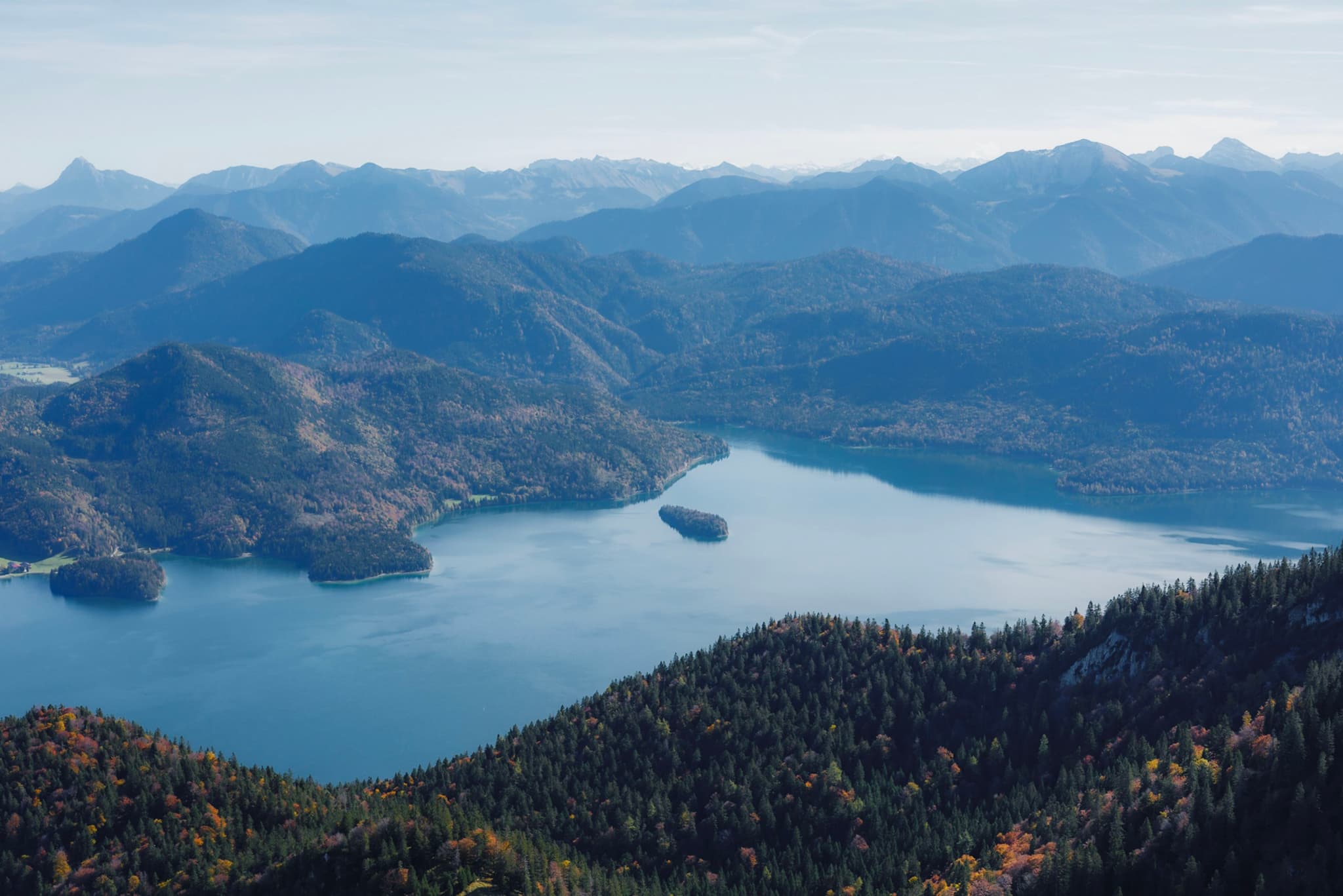 High-altitude view of a large blue lake winding among forested mountains and distant hazy peaks under a clear sky