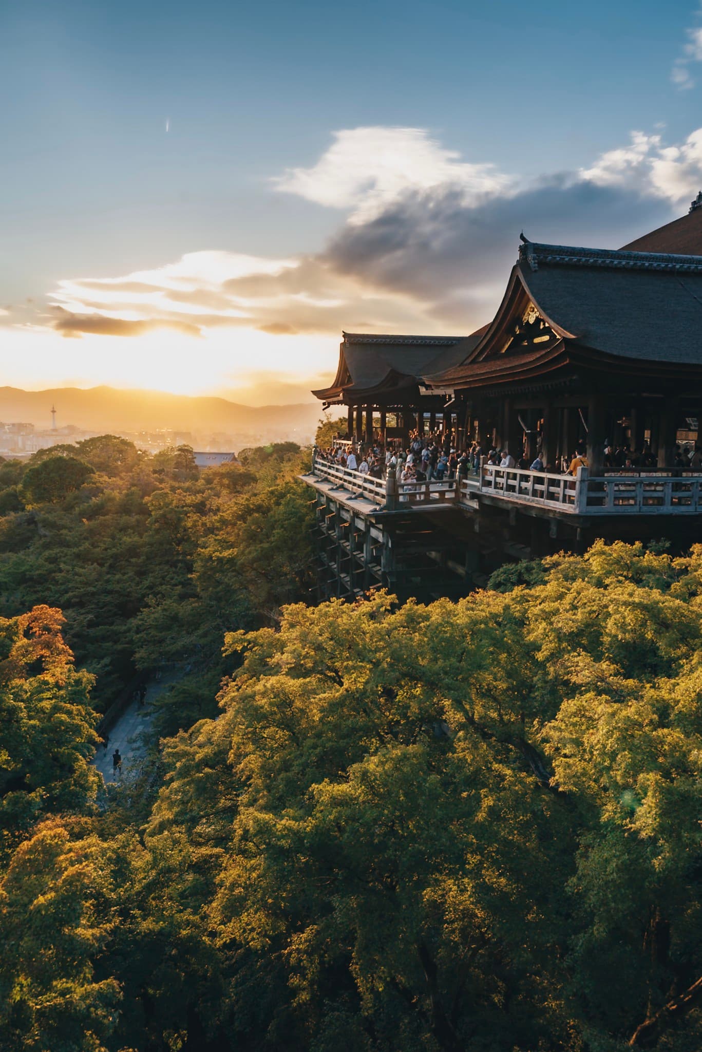 Traditional wooden hillside temple overlooking dense green foliage at sunset with distant mountains and glowing sky
